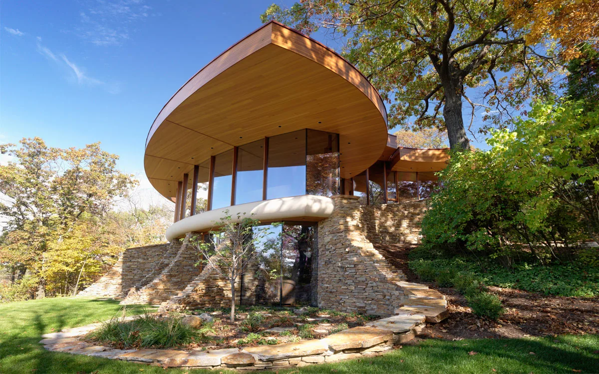 Curved modern house exterior with stone walls, wooden soffit, and floor-to-ceiling windows surrounded by landscaping and trees.