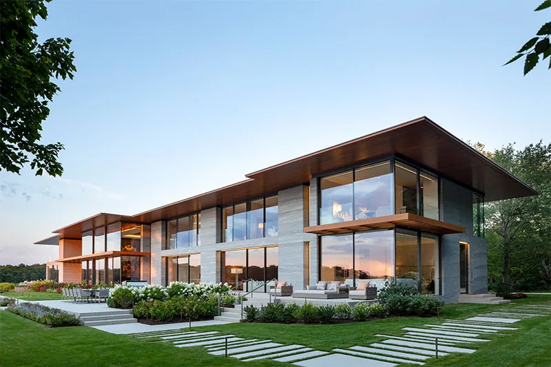 Modern two-story house with extensive floor-to-ceiling glass windows and stone exterior walls during sunset.