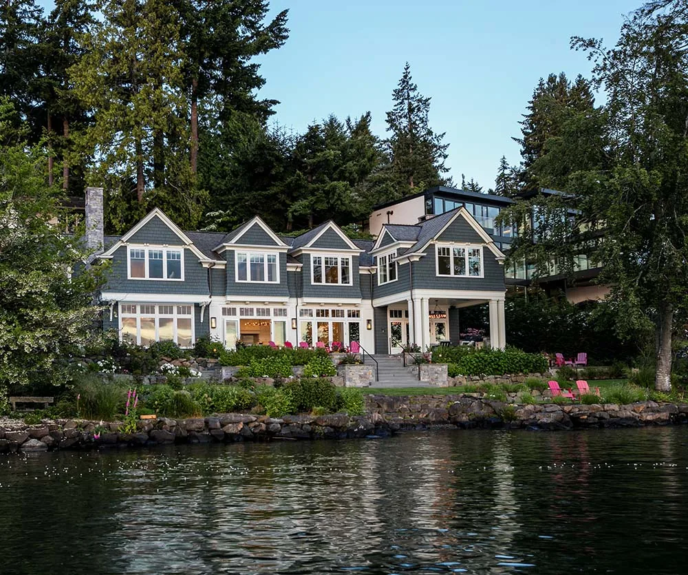 View of a blue-gray house with white trim and multiple gable roofs facing a water body, surrounded by trees.