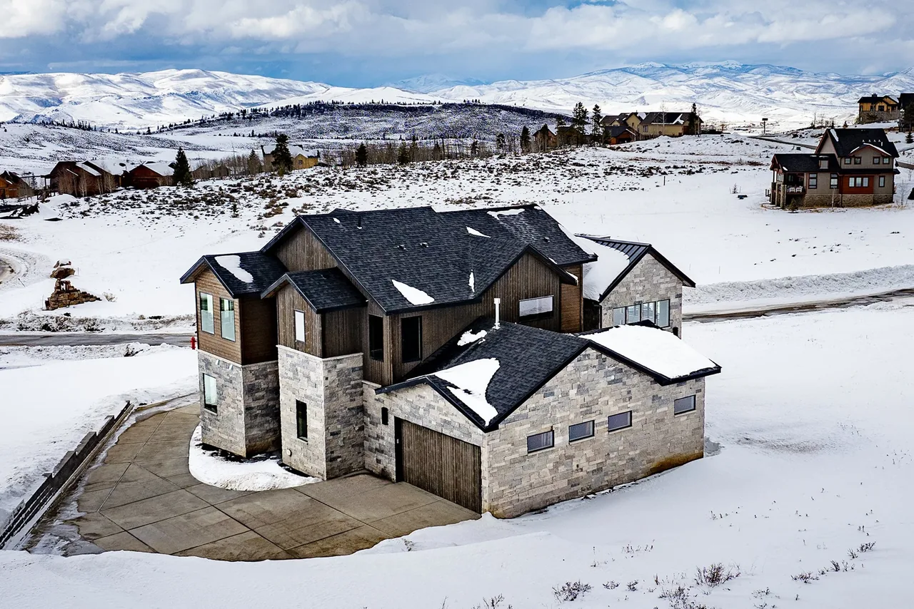 A multi-level house with stone and wood siding sits in a snowy landscape with mountains in the background.
