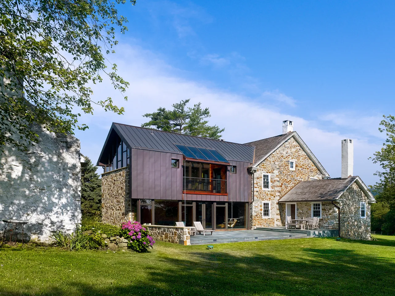 A stone house with a new dark metal addition featuring a pitched roof and solar panels, set in a green yard under a blue sky.