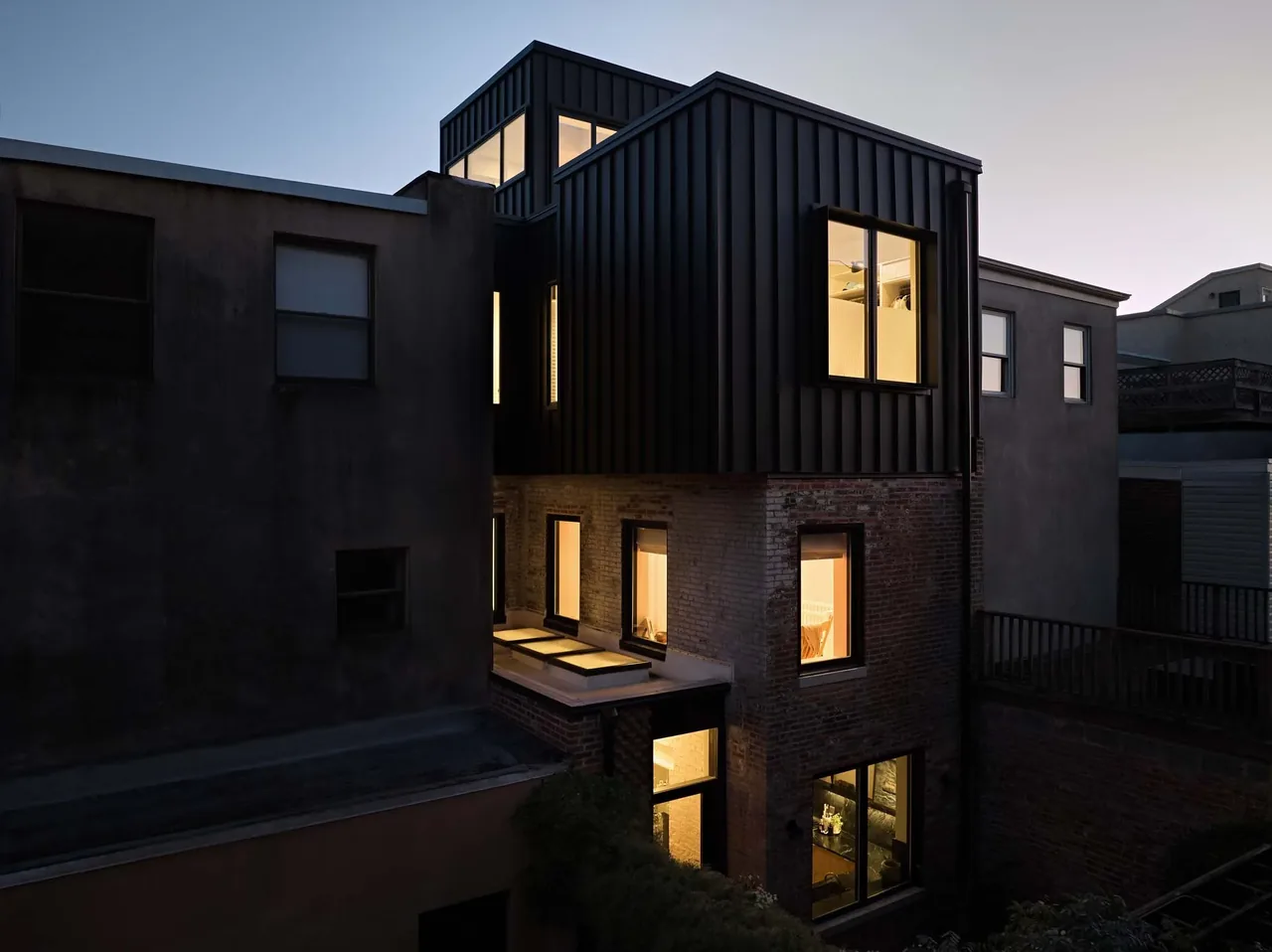 Exterior view of Two Door House at dusk showing a modern three-story building with black metal cladding over red brick base and illuminated,