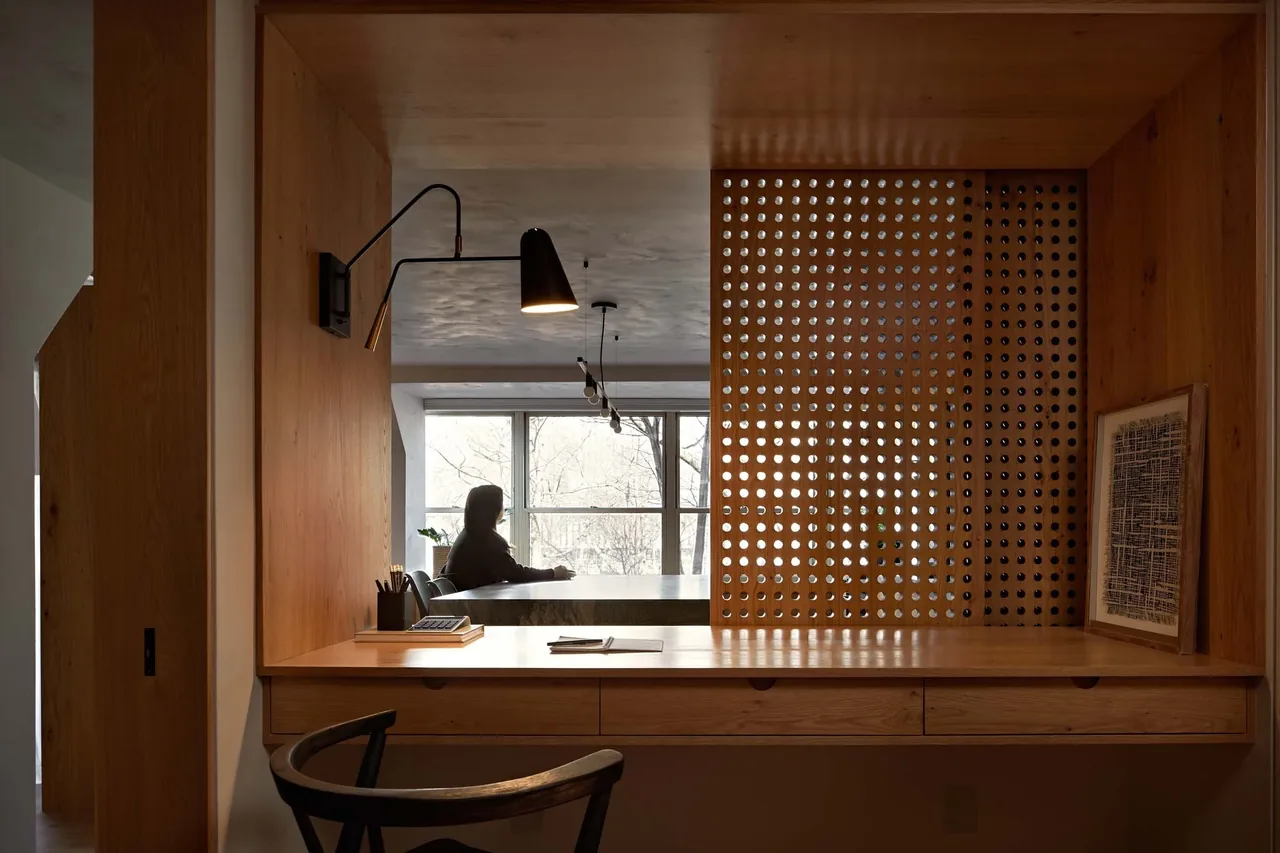 A wooden desk area with perforated paneling and a black wall lamp, overlooking a window with a person sitting at a table beyond.