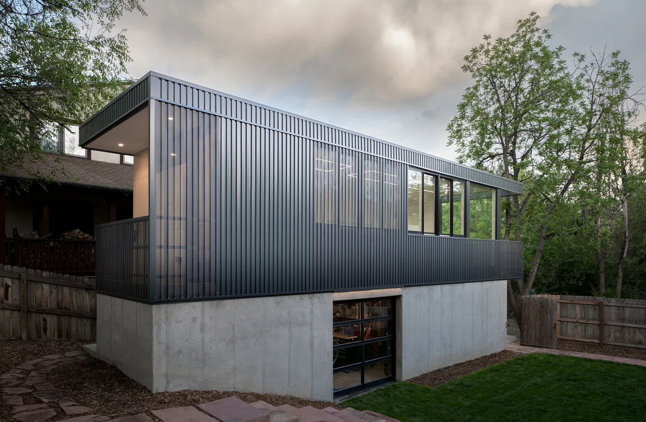 A modern two-level building with a concrete lower level and a dark vertical metal panel-clad upper level with windows, set in a yard with a,