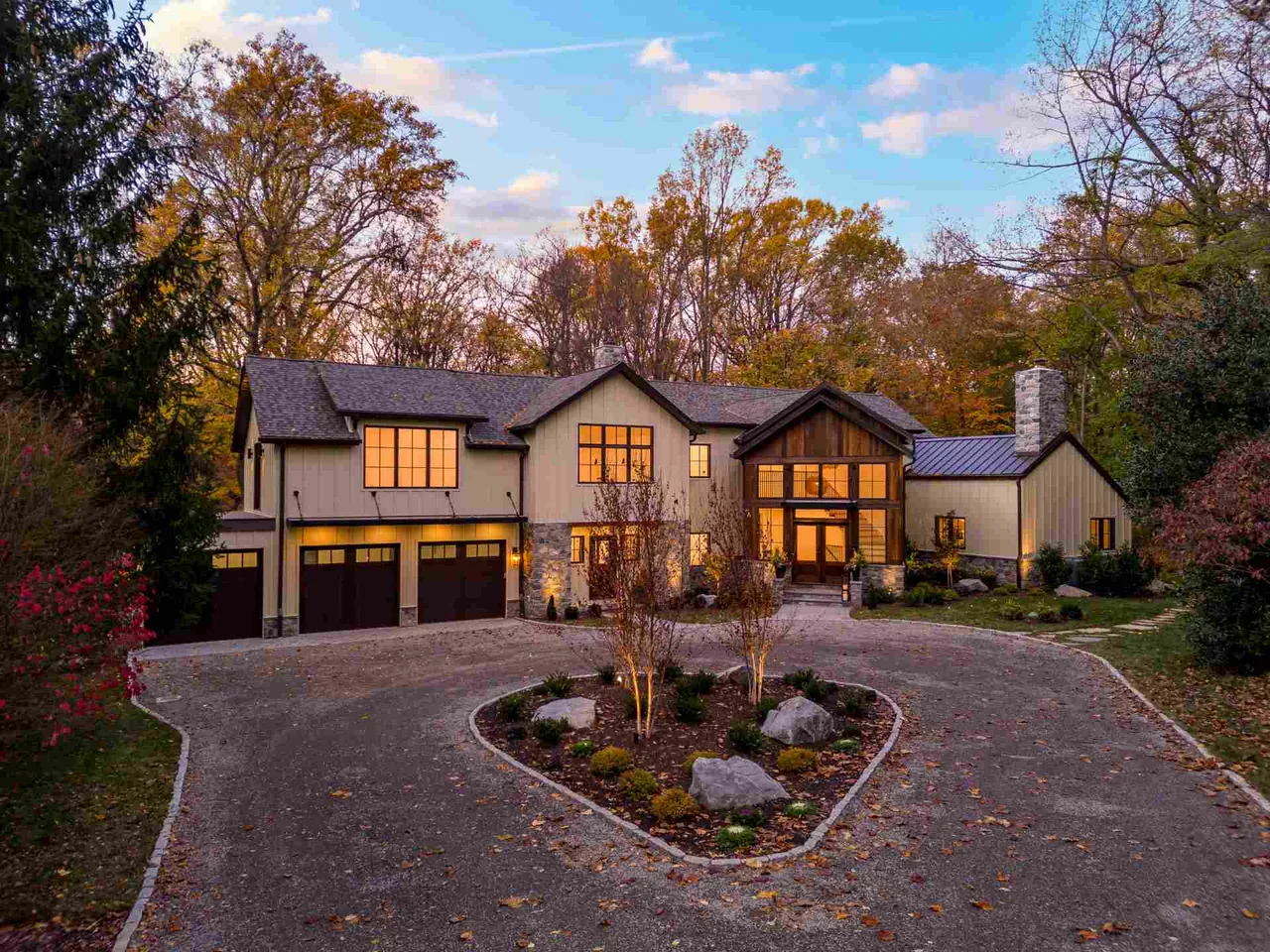 Front exterior view of a two-story house with large windows and a three-car garage at dusk surrounded by trees.