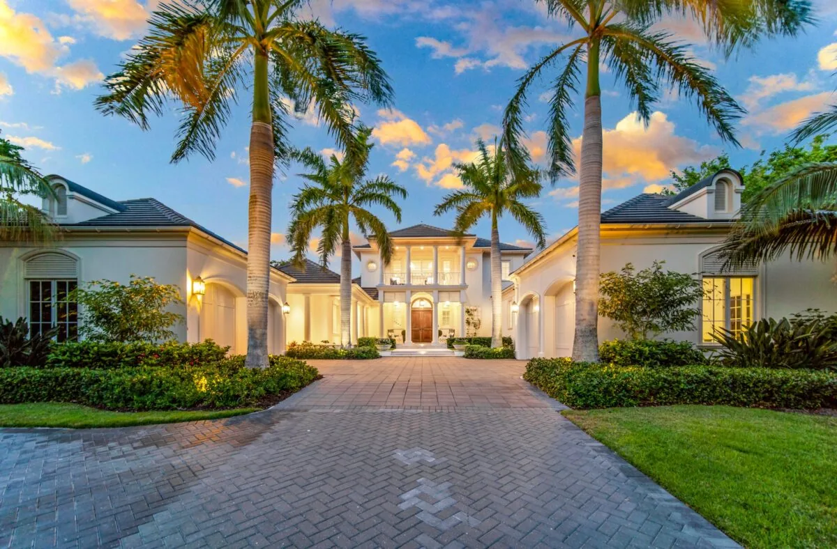 Front exterior of a white bayfront residence with tile roof, columns and palm trees at sunset.