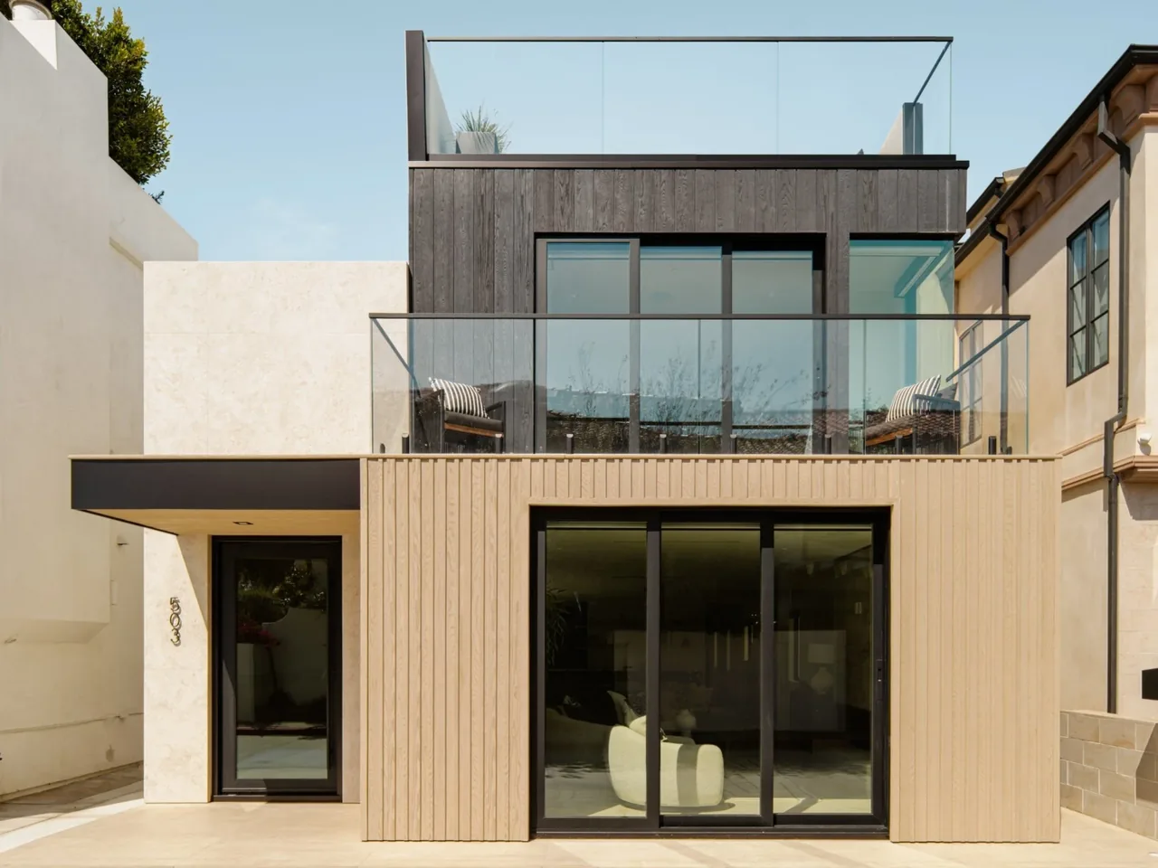 Modern two-story house exterior with beige and dark wood siding, large black-framed windows, and glass balcony railing.