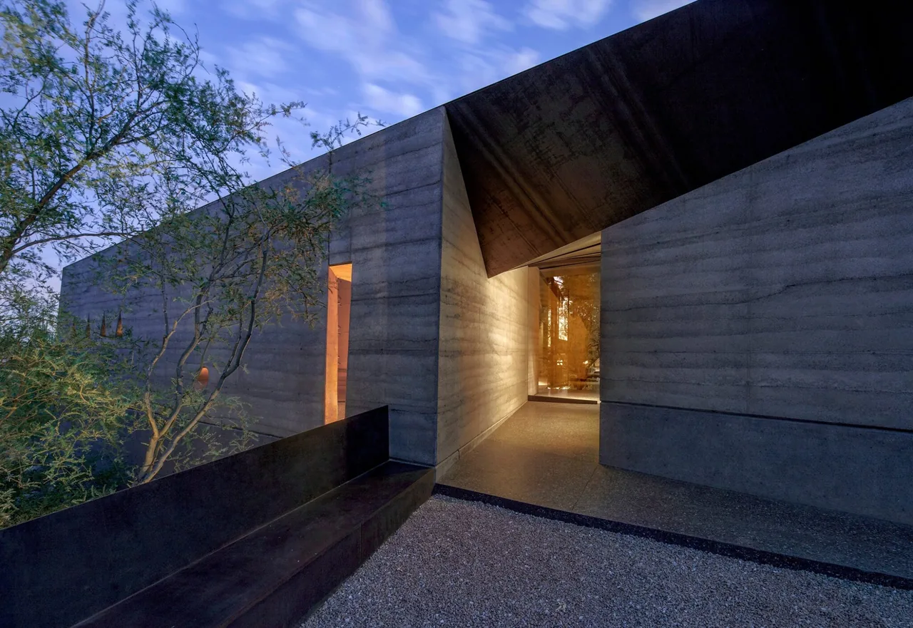 Entrance corner of a modern concrete house with a narrow pathway and integrated bench at dusk.