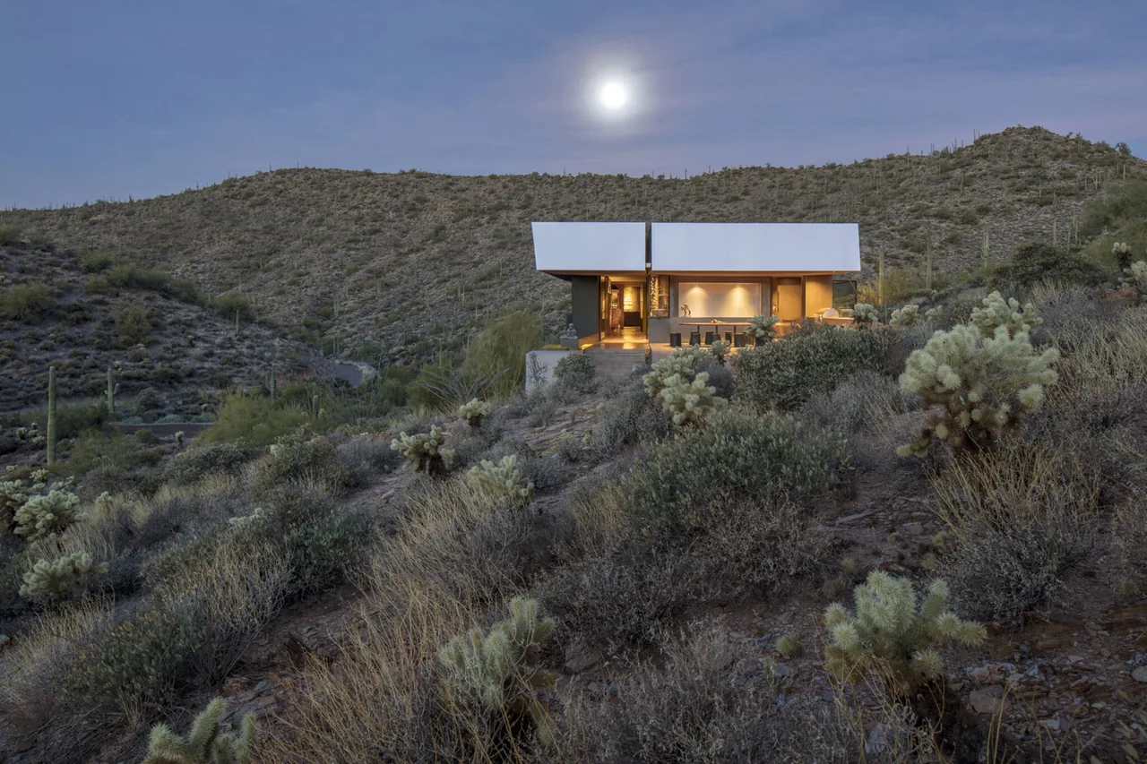 Modern house with illuminated interior in a desert landscape at dusk under a full moon.