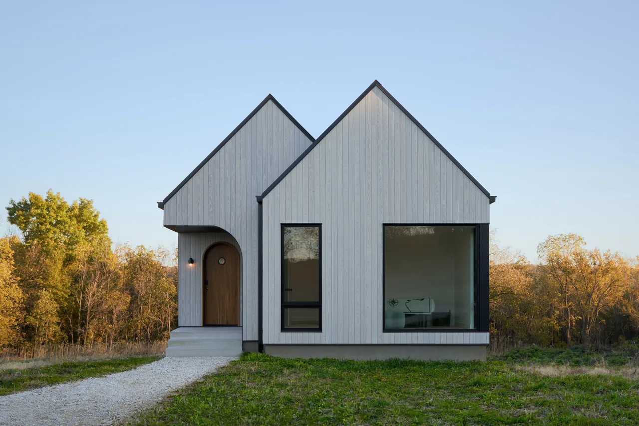Front view of a modern cabin with twin gable roofs, light vertical wood siding, and a large window showing an interior piano.