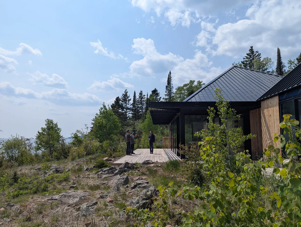 A modern cabin with black metal roof and wooden siding overlooking a rocky and tree-filled landscape with three people standing on the deck.