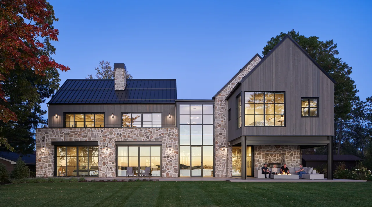 Exterior view of a modern lakehouse with stone and wood siding, large windows, and a covered patio with people sitting around a fire pit.