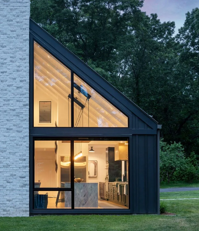 Evening view of a modern house exterior showing large black-framed windows looking into a kitchen with high vaulted ceiling.