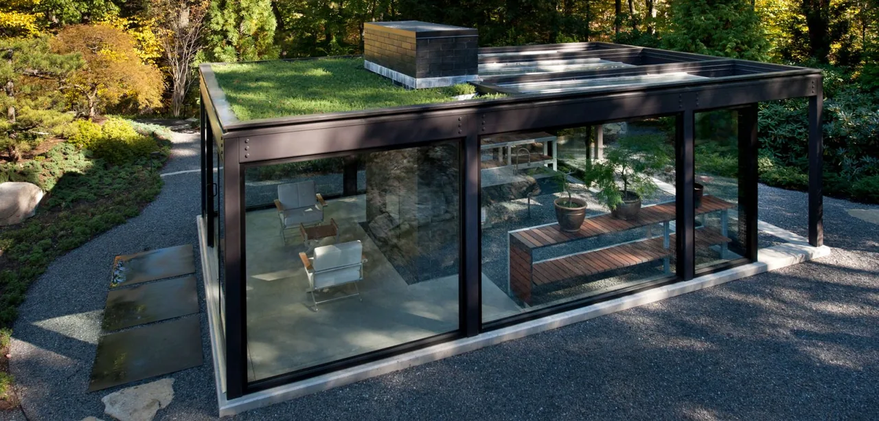 A modern greenhouse with black metal framing, floor-to-ceiling glass walls, and a green roof surrounded by trees.