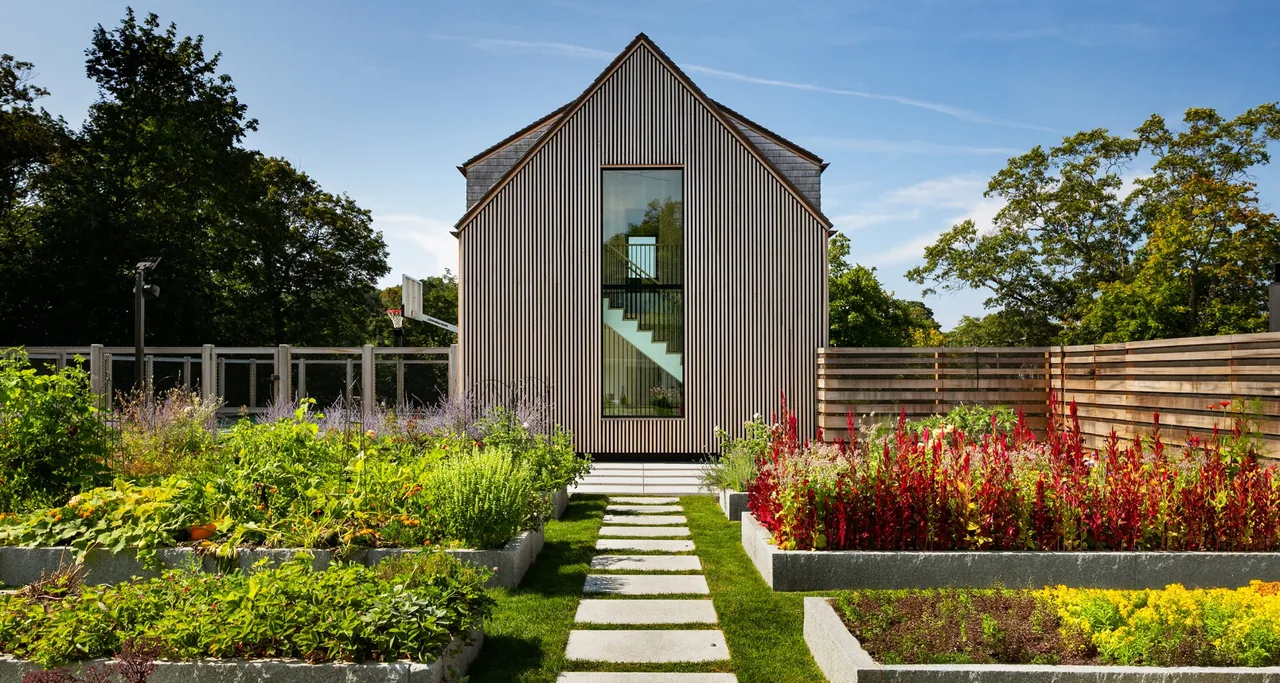 A narrow modern building with vertical siding and a large window revealing an interior stair, surrounded by raised garden beds and a pathway