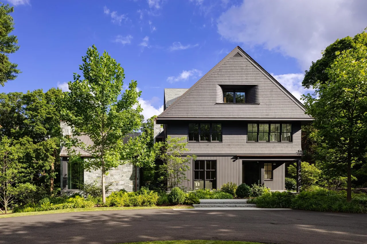 Front exterior view of a dark gray carriage house with a steep gable roof and surrounded by trees.