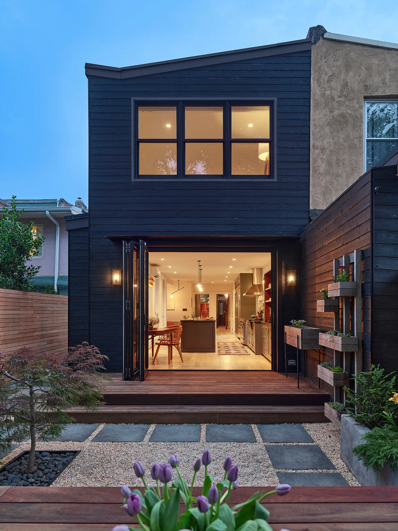 Evening view of a modern black wood-sided two-story house with large windows and an open folding glass door leading to a kitchen with a wood