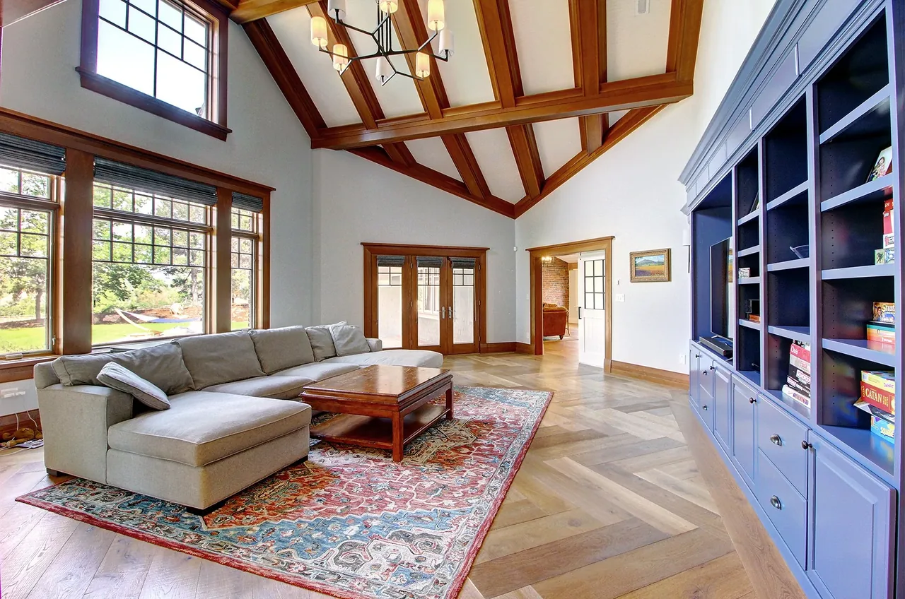 Living room with high vaulted ceiling featuring exposed wooden beams, large windows, sectional sofa, and a blue built-in bookshelf.