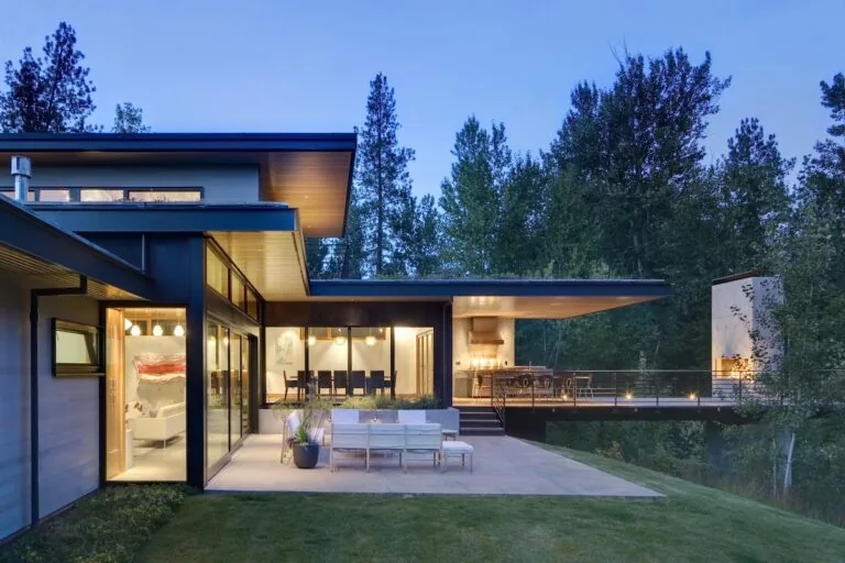 Modern house exterior at dusk with large windows, outdoor seating, and a patio surrounded by trees.