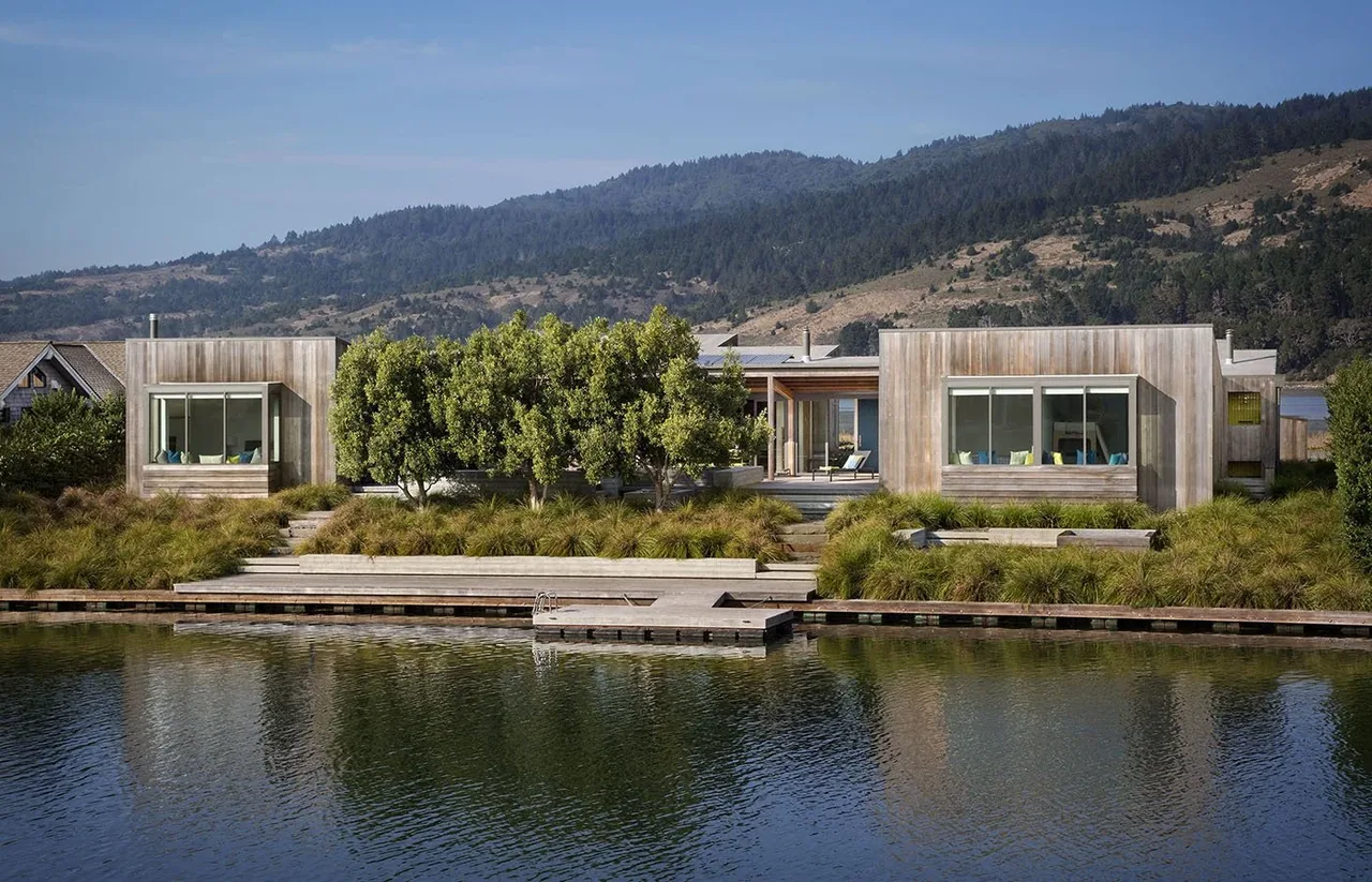 Modern house with wood siding situated next to a calm lagoon with mountainous landscape in the background.