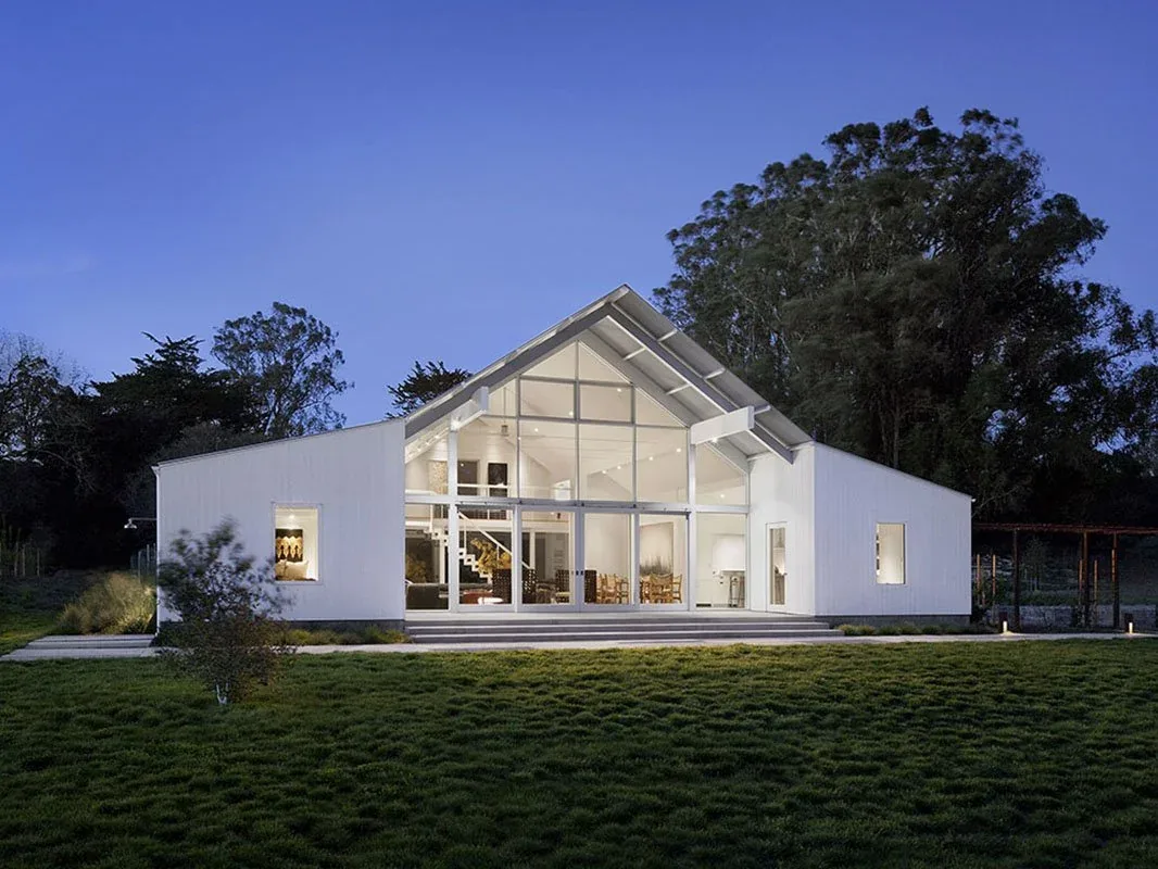 White modern house with large glass facade and a gable roof during twilight.