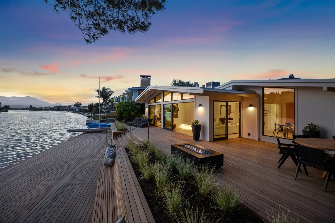 Modern waterfront home with large wooden deck and fire pit at sunset.