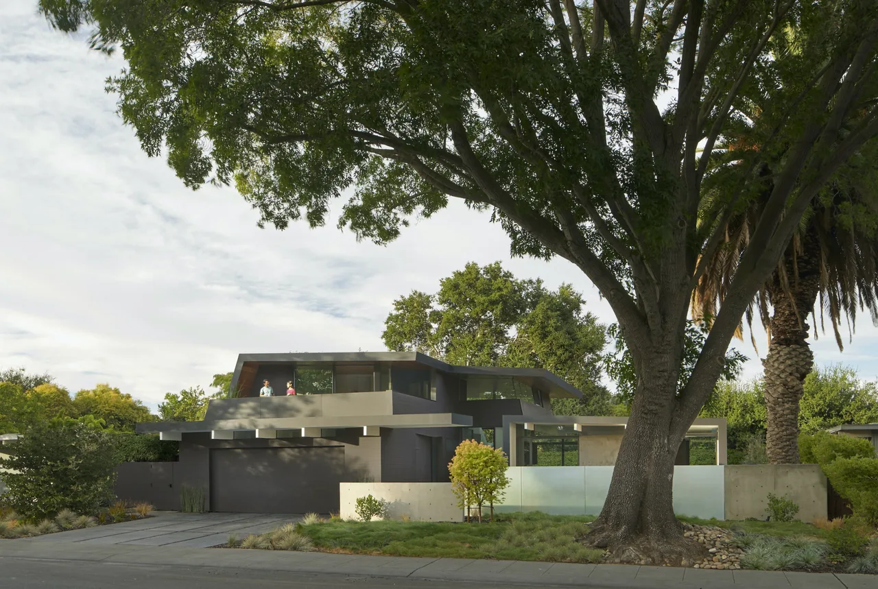 Modern two-story gray house with flat roof and large trees in the front yard.