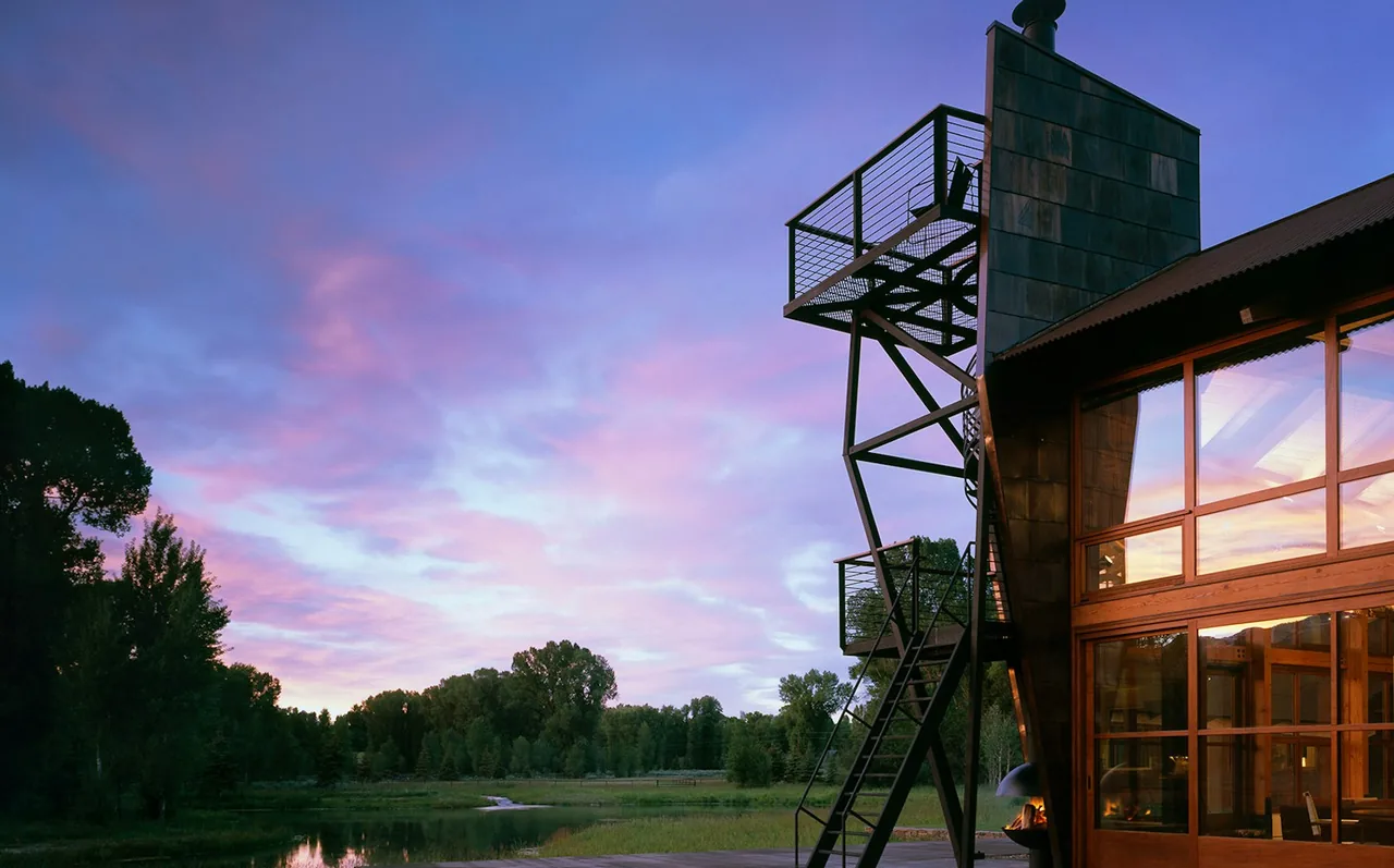 Evening view of a modern home exterior with large windows and a metal staircase tower beside a serene water landscape.