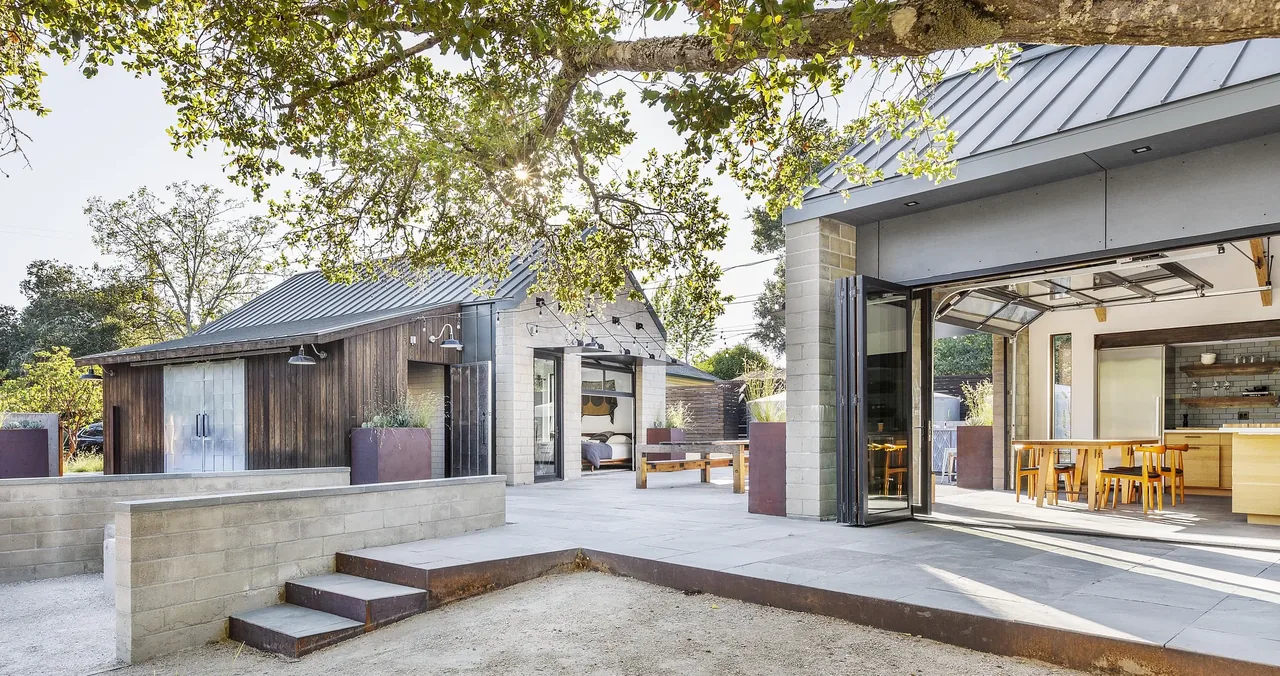 Outdoor view of a modern farmhouse-style home with metal roofs, wood siding, and an open kitchen-dining area visible through large folding,玻