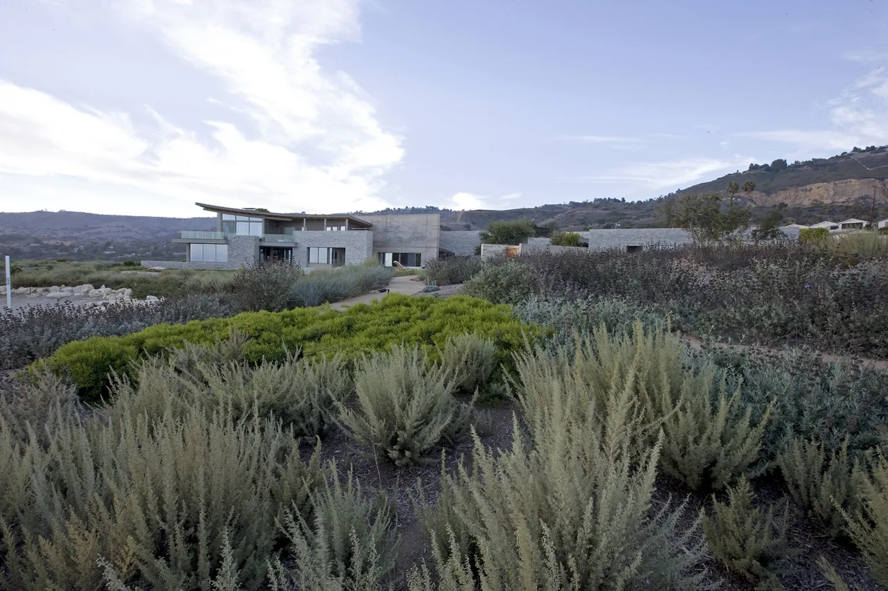 Modern house with stone exterior walls set in a dry landscape with shrubs and hills in the background.