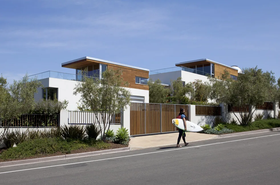 Street view of a modern two-story house with wood and white stucco exterior and large windows behind a gated fence with greenery.
