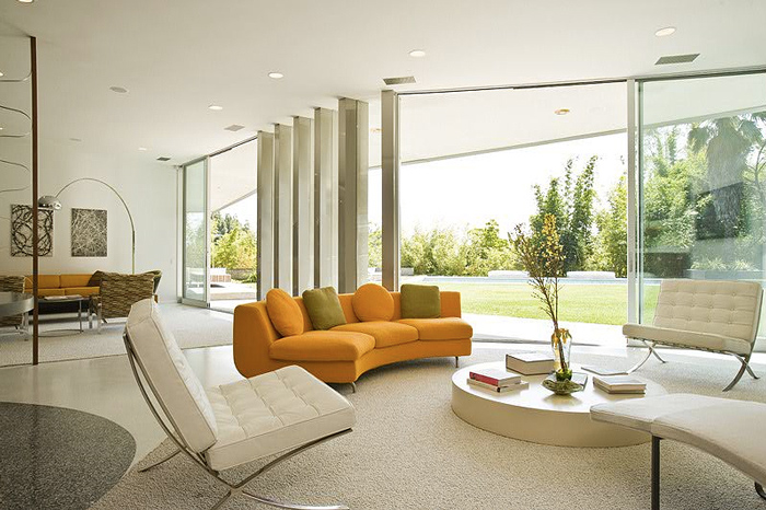 Modern living room with white leather chairs, an orange sectional sofa, and floor-to-ceiling windows looking out to a green lawn.