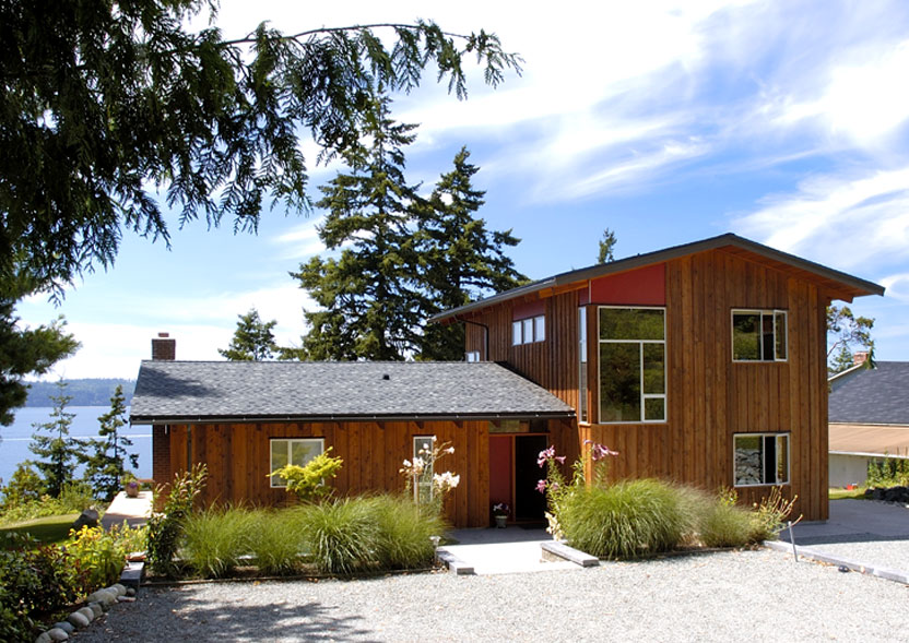 Wood-paneled two-story house with large windows and a gravel driveway surrounded by greenery and trees.