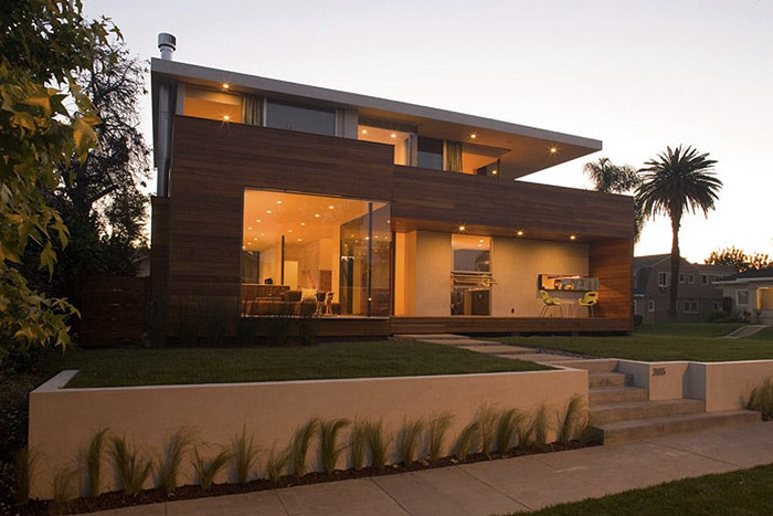 Evening view of the rear exterior of a modern two-story residence with large glass windows and wood siding, showing outdoor lighting and a w