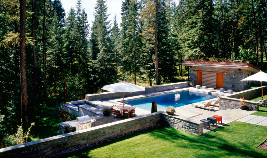 A rectangular outdoor pool set in a natural forested landscape with stone walls and seating areas under umbrellas.