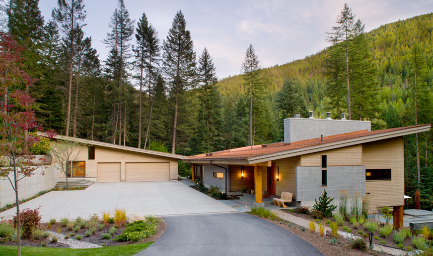 Modern residence featuring sloped roofs, wooden siding, and concrete driveway, surrounded by natural landscaping and tall pine trees.