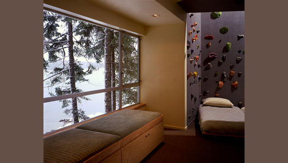Room featuring a cushioned bench under large metal-framed windows with a climbing wall beside a daybed, overlooking a snowy lake view.