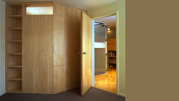 Interior view showing built-in wood cabinetry and an open door to a room with bookshelves and warm wood flooring.