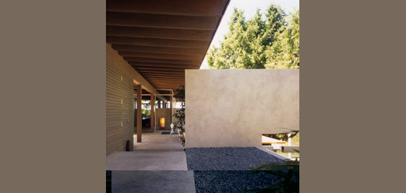 Covered exterior walkway with exposed wood beam ceiling and stucco wall beside gravel ground cover and concrete slabs.