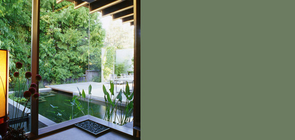 Interior view through floor-to-ceiling glazing to a covered patio and pond with aquatic plants.