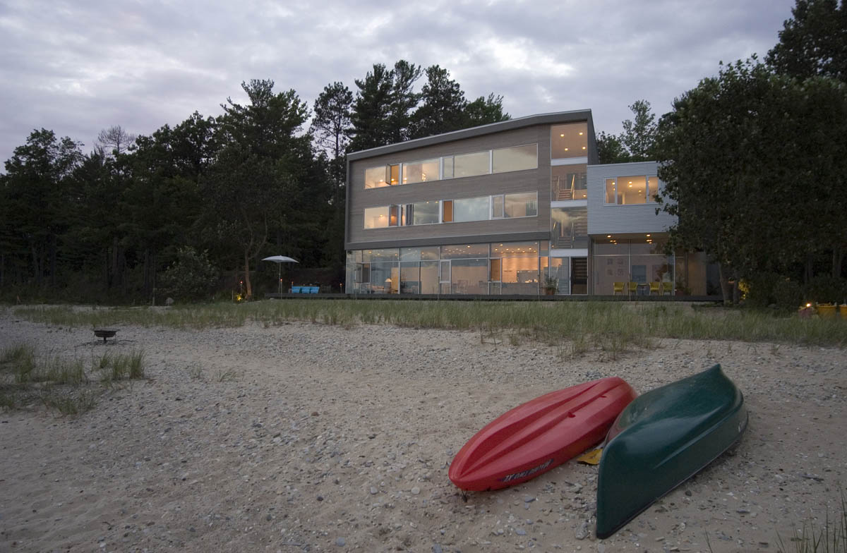 Three-story modern residence with horizontal windows and wood and metal cladding facing sandy waterfront with kayaks in foreground.