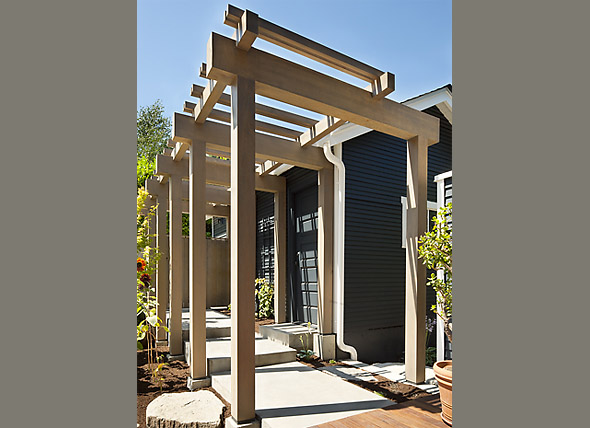 Wooden pergola structure beside a house with dark siding and potted plants in a courtyard setting.