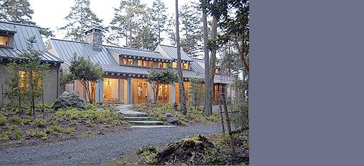 Wood-clad residential building with metal gable roofs and large windows set in wooded landscape.