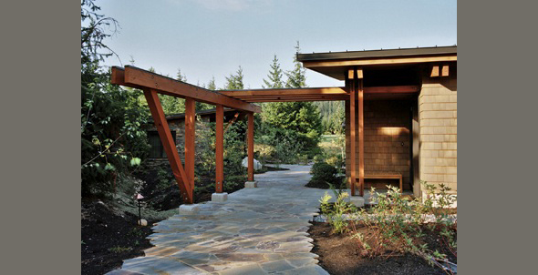 Stone pathway and wooden pergola frame the entry of Mountain Meadow House with shingle siding and sloped roof.