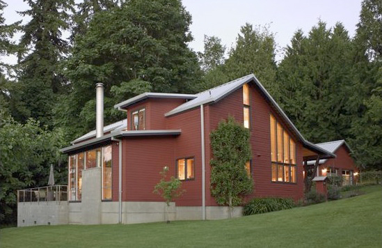 Side view of a red two-story house with large vertical windows, situated on a grassy hill with tall trees in the background.