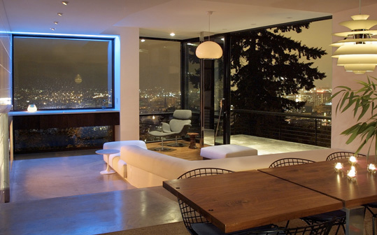 Modern nighttime interior of a living and dining space with corner windows overlooking the city.