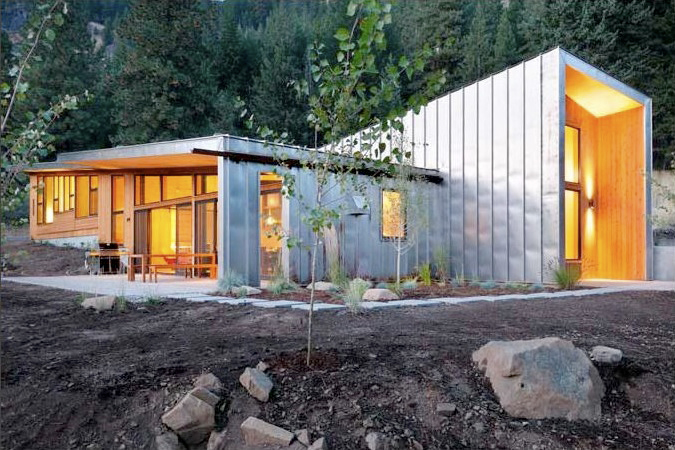 Modern house exterior with standing seam metal siding and large windows illuminated at dusk in a forested area.