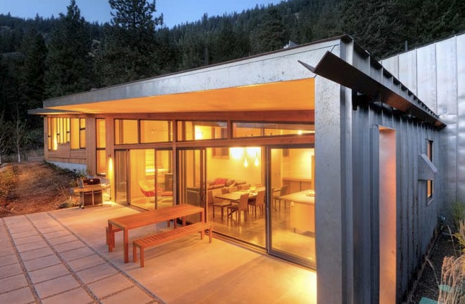 Miner's Refuge features metal siding, large sliding glass doors, and a concrete patio illuminated by warm interior lighting at dusk.