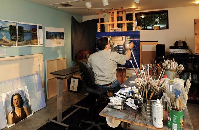 Artist working on a still life painting in a home studio filled with art supplies and paintings.