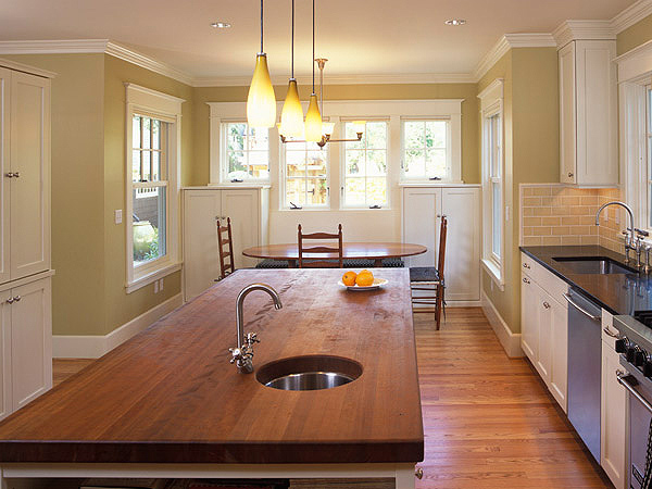 The Cady Residence kitchen features a wooden island with mounted sink and pendant lighting over a dining nook with wood chairs and yellow walls.